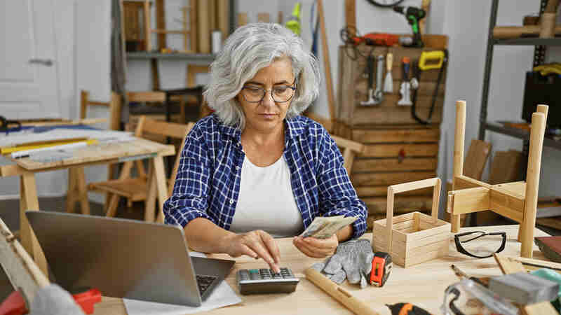 Mature grey-haired woman counting money while using a calculator and a laptop
