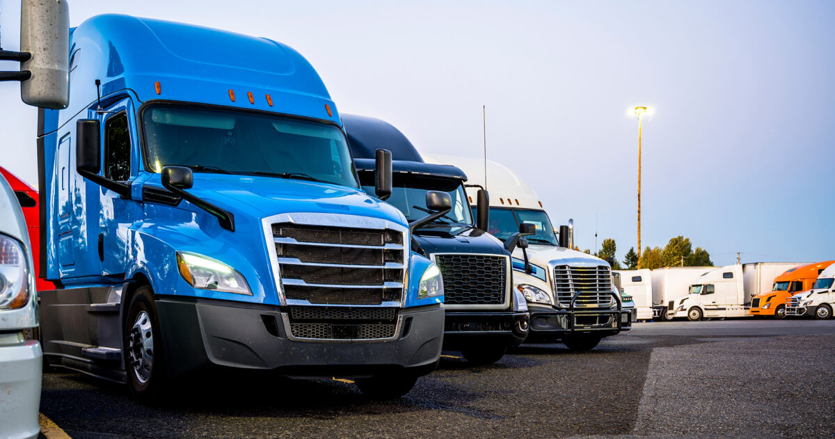 Different-big-rigs-semi-trucks-standing-in-row-on-the-truck-stop-parking-lot-at-evening-time.